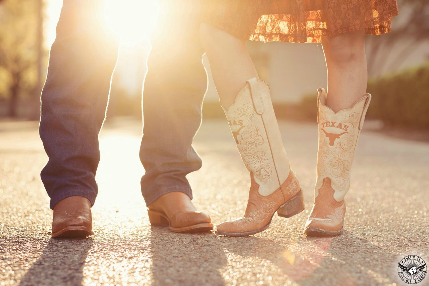 Two people stand in wearing an orange lacey dress and blue jeans with Texas Longhorn boots with Longhorn logo on an exposed aggregate side walk with the warm yellow orange sun shinning through their legs in this fun engagement photo in Austin, Texas.
